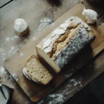 A freshly baked banana bread loaf sliced neatly on a wooden cutting board, surrounded by ripe bananas and a light dusting of flour.